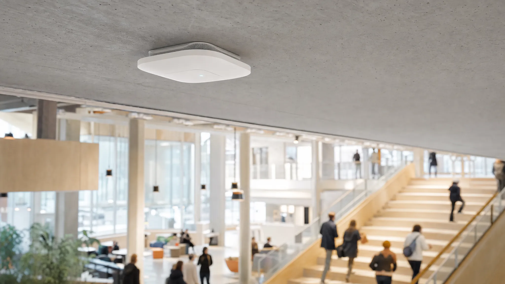 Un punto de acceso Wi-Fi empresarial montado en el techo, sobre personas que se mueven por un espacio público luminoso.