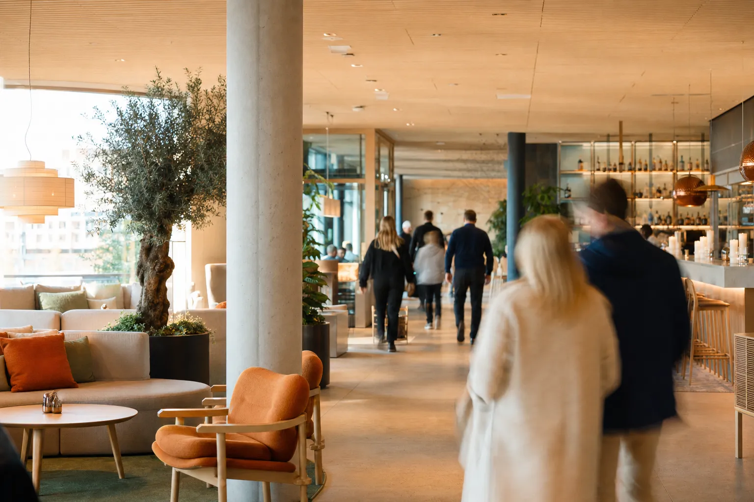 Des clients traversent le lobby et le bar chaleureux d’un hôtel scandinave haut de gamme, vus de dos dans une douce lumière du soir.