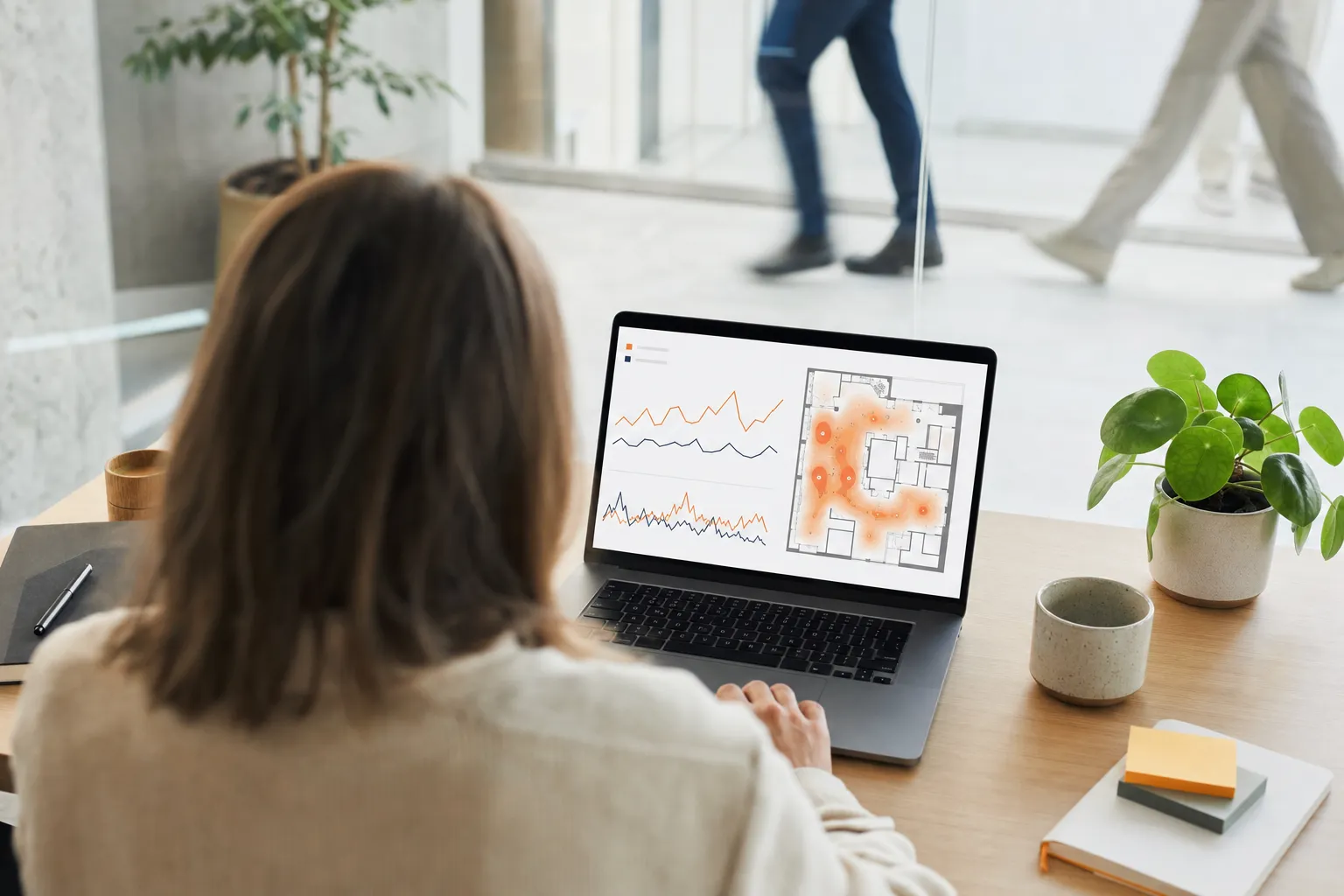 Une analyste examine le tableau de bord Bumbee Labs sur un ordinateur portable, des graphiques de tendances de fréquentation à côté d’une carte de chaleur orange sur le plan d’étage d’un site, avec des personnes qui passent en arrière-plan.
