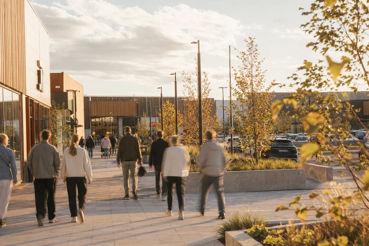 Besökare som går mellan stora butiker över ett öppet torg på en extern handelsplats i kvällssol, sedda bakifrån.