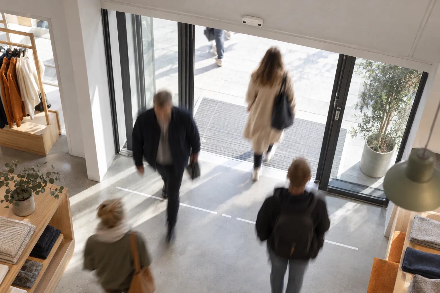 Sett fra innsiden av en butikkinngang: en takmontert optisk 3D-sensor teller kunder som krysser en inn/ut-linje, i bevegelsesuskarphet uten gjenkjennbare ansikter.