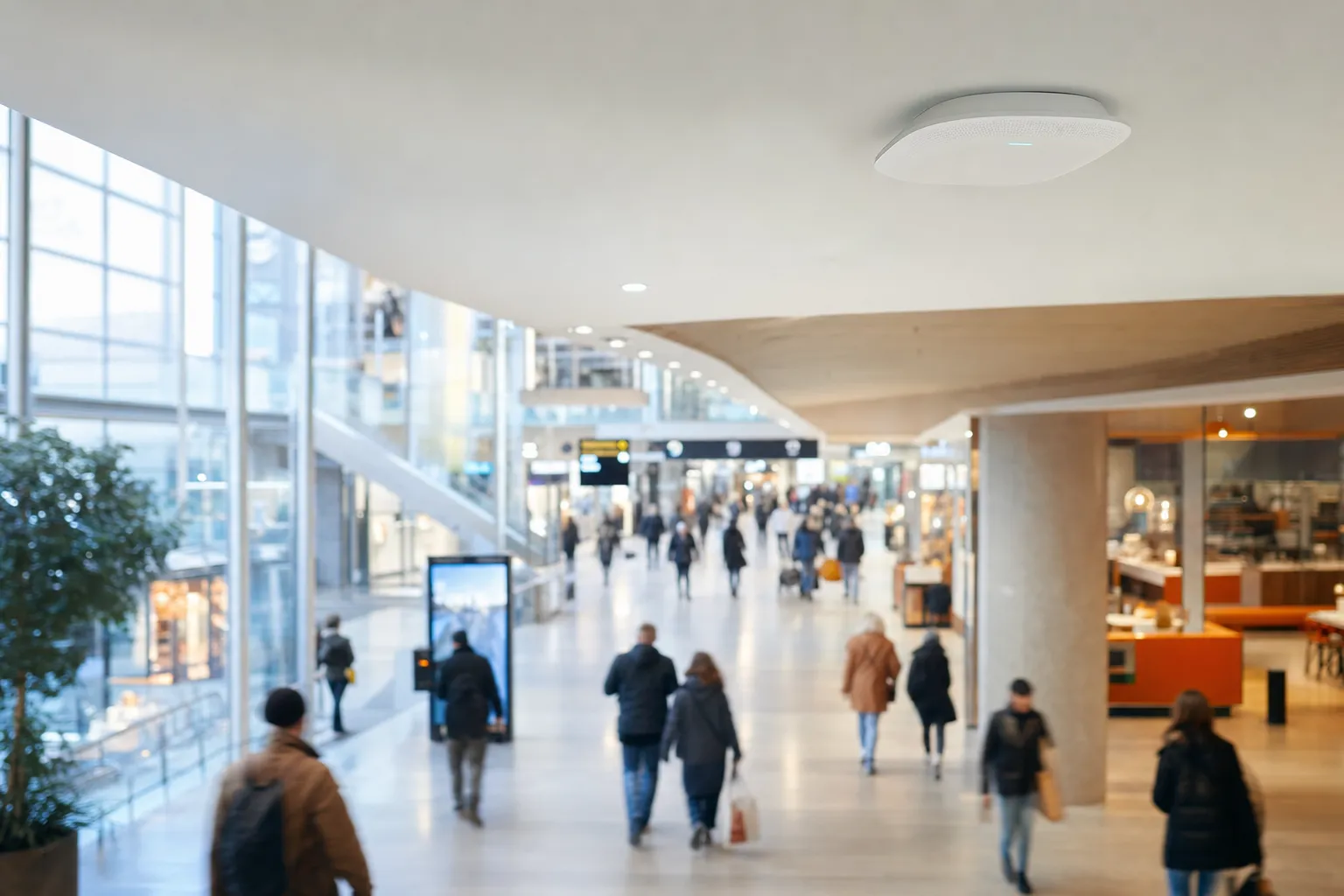 Un access point Wi-Fi a soffitto sopra i visitatori in movimento in un centro commerciale luminoso, con effetto mosso.